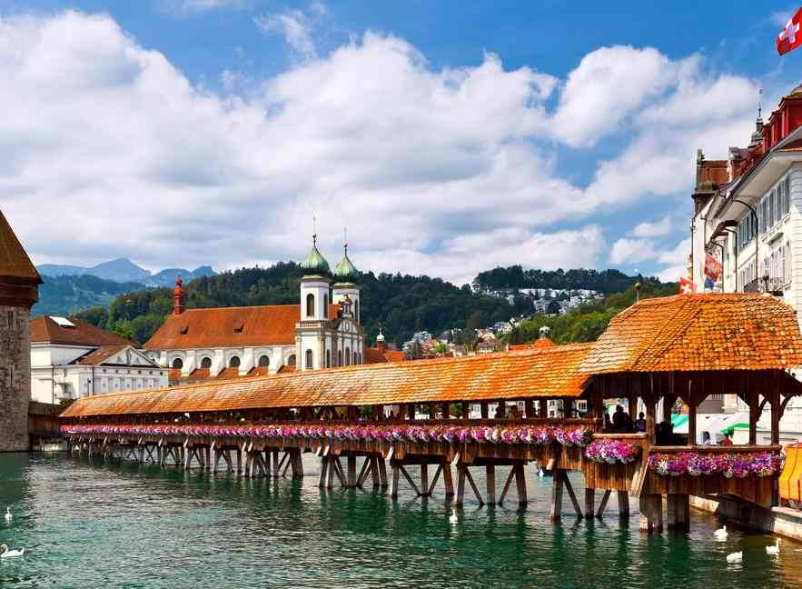 Covered wooden bridge decorated with flowers crossing a river in Lucerne, Switzerland, with historical buildings and mountains in the background under a partly cloudy sky.